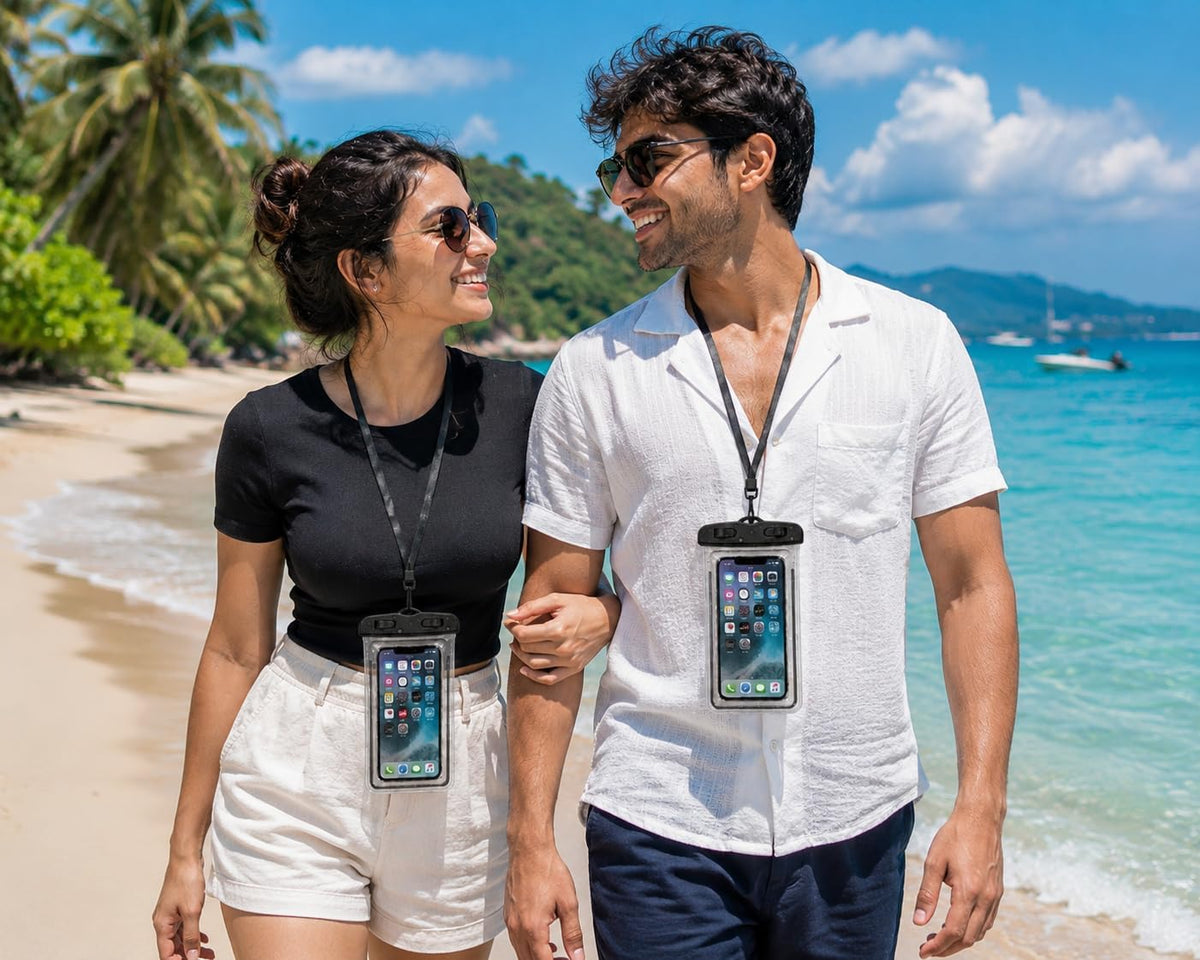 A man and a woman walking arm-in-arm on a sunny beach, both wearing black waterproof phone pouches hanging from lanyards around their necks. The pouches securely hold smartphones, with the woman's pouch showing a black case and the man's showing a clear case with a black strap.