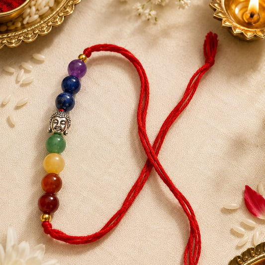 A red cord bracelet with a sequence of multicolor beads (purple, blue, green, yellow, orange, red) and a silver Buddha head charm, styled on a beige fabric with flowers, rice, and a diya.