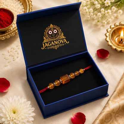 A beaded Rakhi with amber-colored beads and a red cord rests inside an open dark blue presentation box. The box lid features a black insert with a gold Jaganova logo.