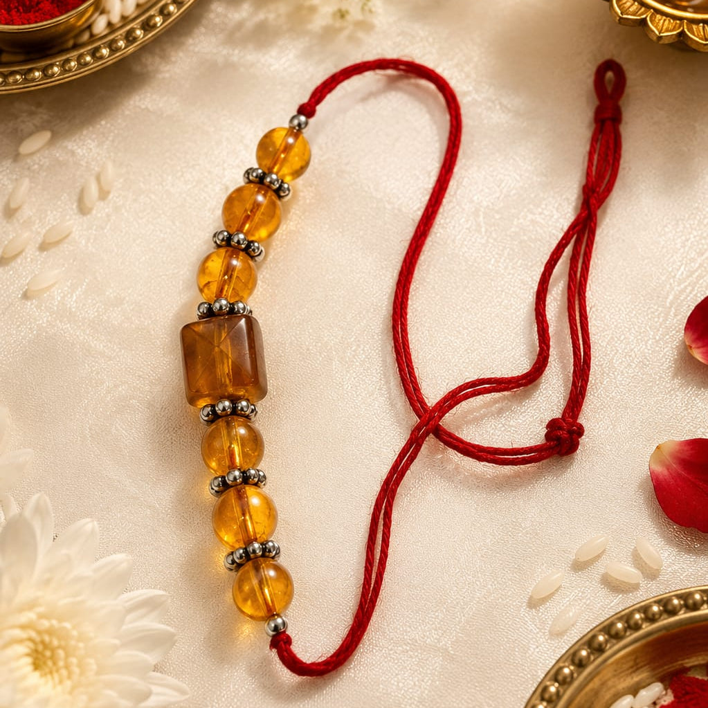 A traditional Indian Rakhi beaded wrist strap with translucent amber-colored beads and a red cord, displayed on a cream fabric background with festive props like a brass diya, rice, and flower petals.