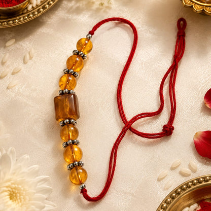 A traditional Indian Rakhi beaded wrist strap with translucent amber-colored beads and a red cord, displayed on a cream fabric background with festive props like a brass diya, rice, and flower petals.