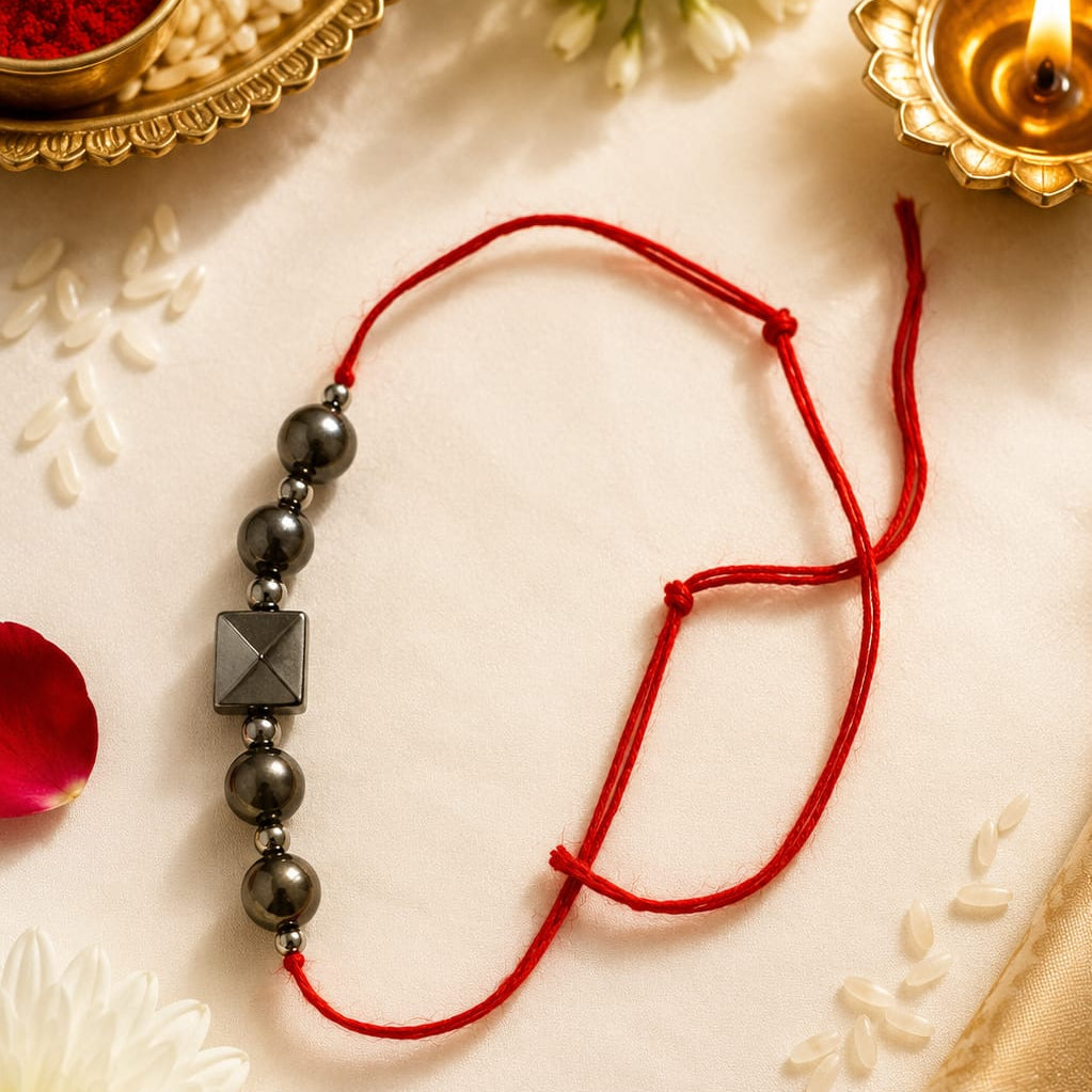 A red string rakhi bracelet with dark metallic beads and a central pyramid-shaped charm, styled on a cream fabric with flowers, a diya, and a bowl of rice.
