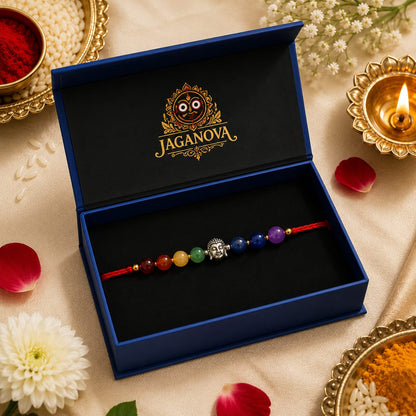 A beaded Krishna rakhi bracelet displayed inside an open blue Jaganova gift box, surrounded by festive props including a lit diya, flowers, and red petals on a beige fabric background.