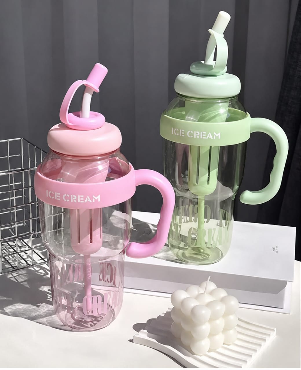 Two clear plastic handled drinking cups with integrated straws and ice cube holders, one in pink and one in mint green, labeled 'ICE CREAM', displayed on a white surface with a gray curtain background.