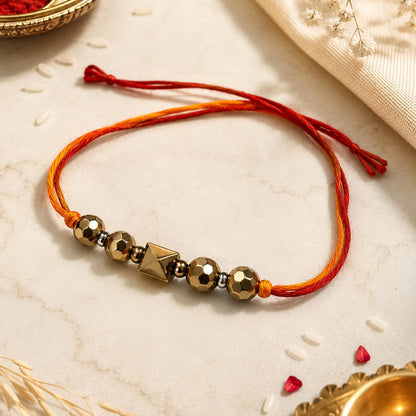 A traditional Indian beaded bracelet (rakhi) made of red and orange threads with metallic gold beads, displayed on a light marble surface with festive props like rice, dried flowers, and brass oil lamps.